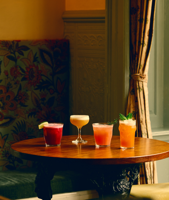 A cozy corner with a round table featuring four colorful cocktails, a floral cushioned chair, and warm window curtains.