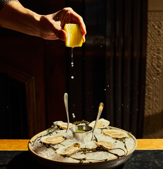 A hand squeezes lemon over a plate of oysters with ice and a small dipping bowl.