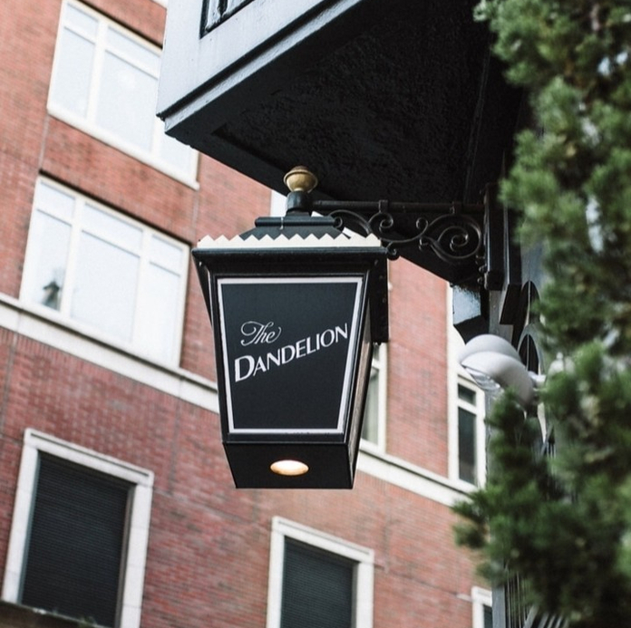 The Dandelion restaurant signboard hanging above a brick building with a window and greenery partially visible.