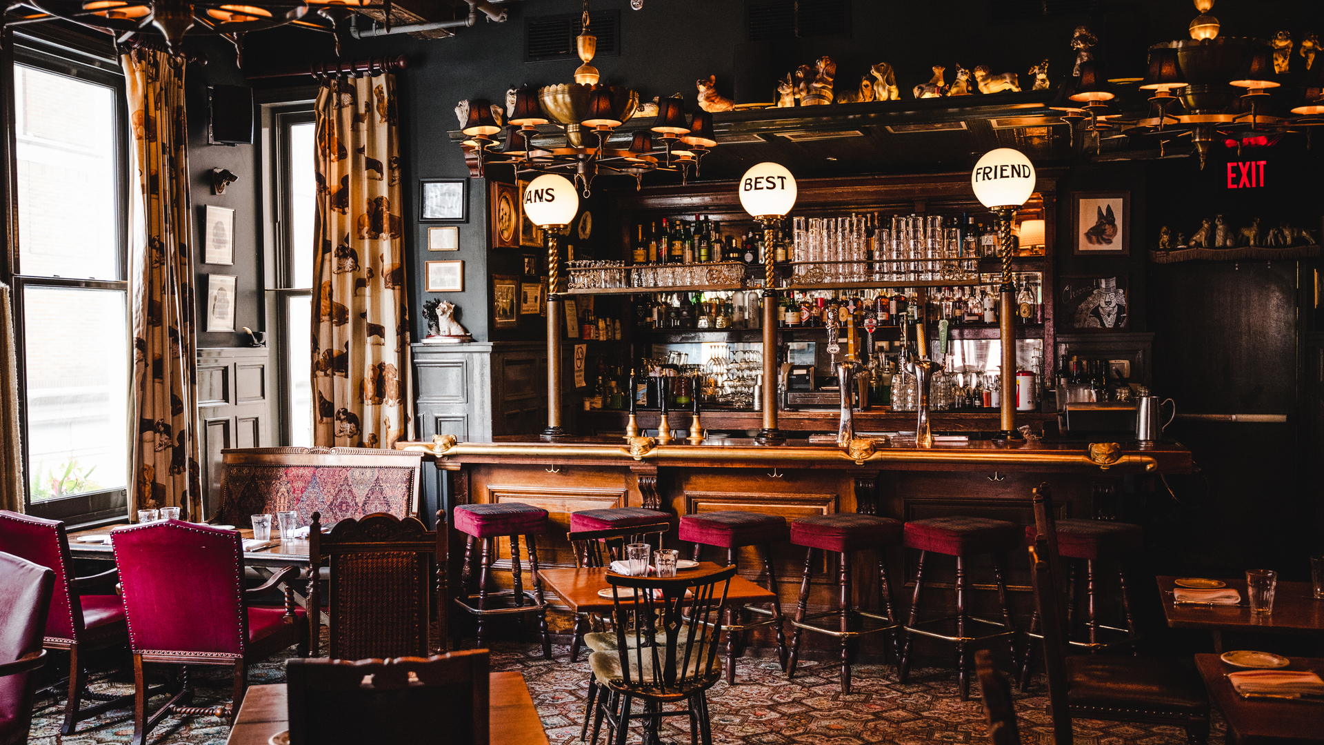Cozy restaurant with dark walls, wooden bar, stools, red chairs, and a large window. Barware and decor visible.