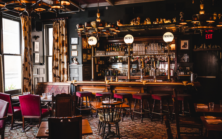 Cozy, dimly lit bar with wooden furniture, patterned carpet, and large windows with curtains. Bar counter has round signs, shelves, and various bottles.