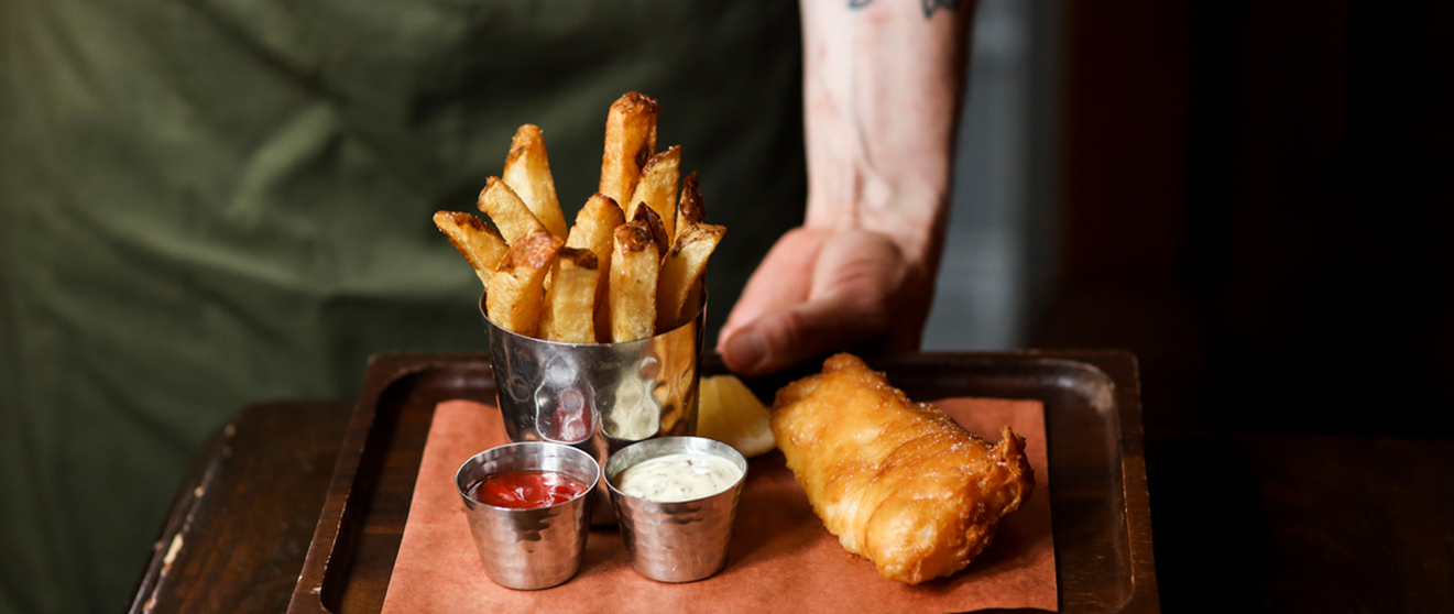 Battered fish and fries with two dipping sauces on a wooden tray, held by a person wearing a green apron.