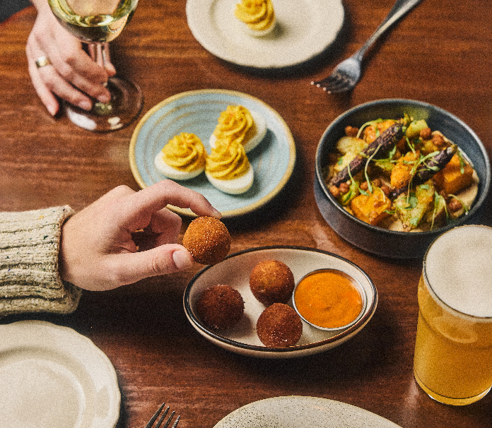 A tabletop spread of small plates and drinks, including deviled eggs, fried bites with dipping sauce, a vegetable dish, toast, a glass of white wine, and a beer.