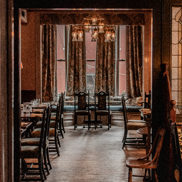 A warmly lit dining room with dark wood trim, set tables, vintage chairs, patterned curtains, and a chandelier centered near a window alcove.