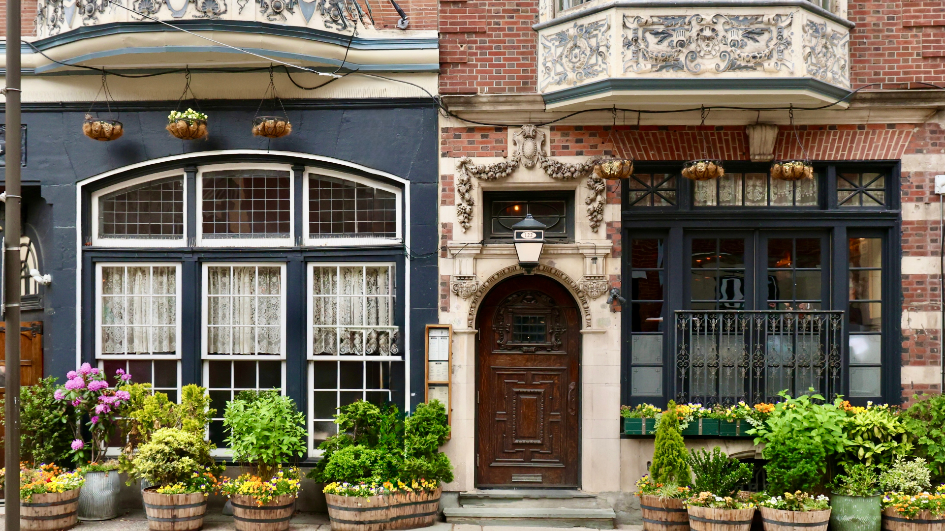 A historic entrance with an ornate wooden door, carved stone archway, hanging planters, and barrel planters filled with greenery along the sidewalk.
