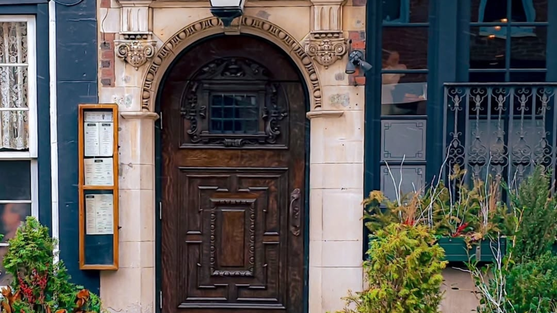 A historic entrance with an ornate wooden door, carved stone archway, hanging planters, and barrel planters filled with greenery along the sidewalk.