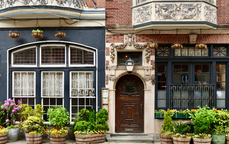 A historic entrance with an ornate wooden door, carved stone archway, hanging planters, and barrel planters filled with greenery along the sidewalk.