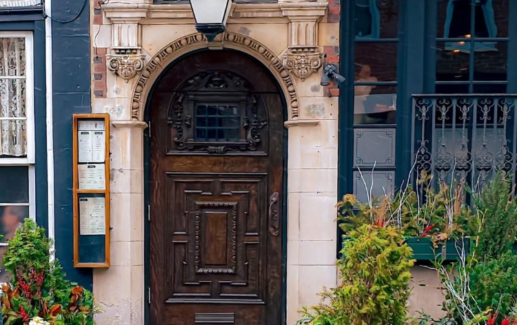 A historic entrance with an ornate wooden door, carved stone archway, hanging planters, and barrel planters filled with greenery along the sidewalk.